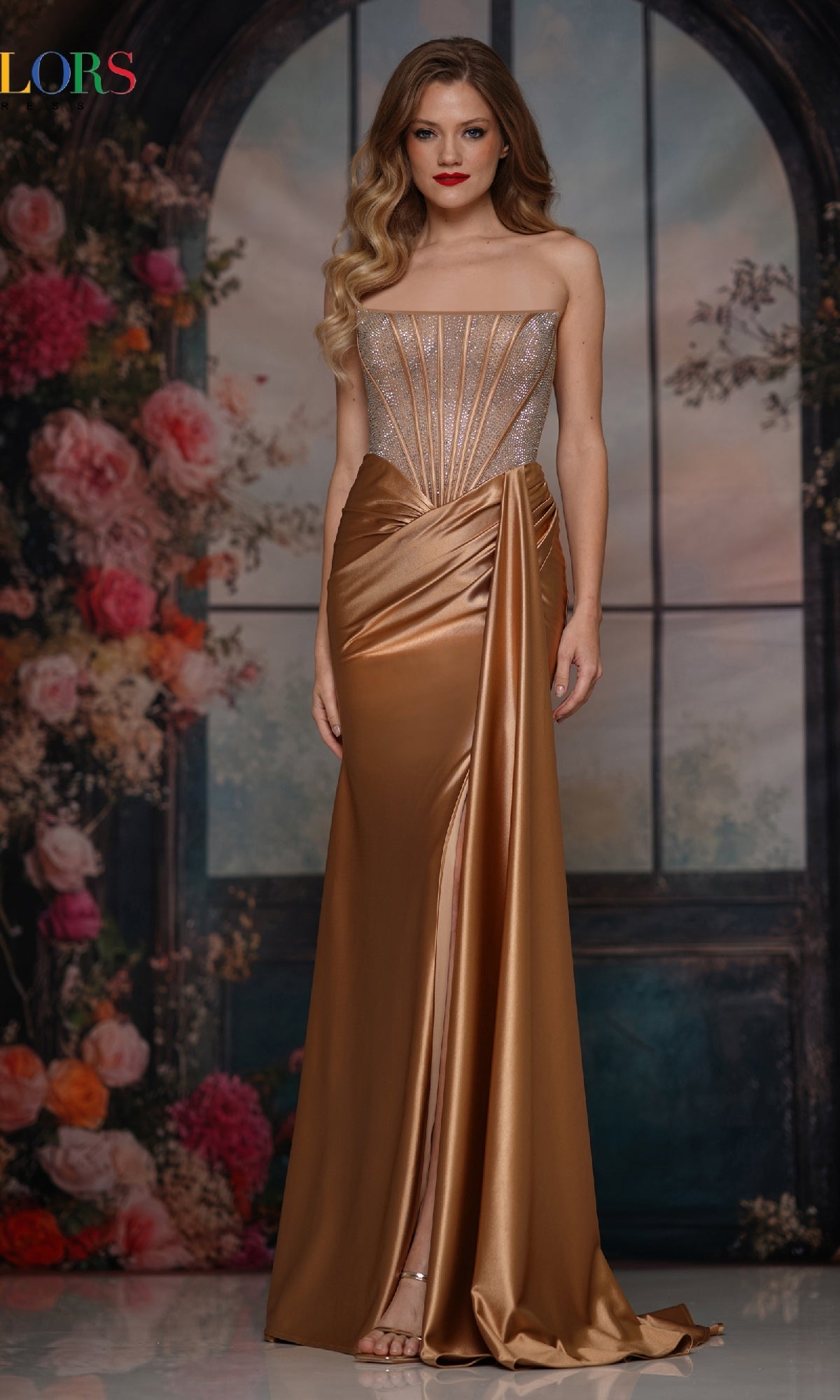 A woman stands before a floral arched window in the Colors Dress 3933 Strapless Long Corset Prom Gown, featuring an embellished corset and a shimmering metallic gold satin skirt with a dramatic high slit.